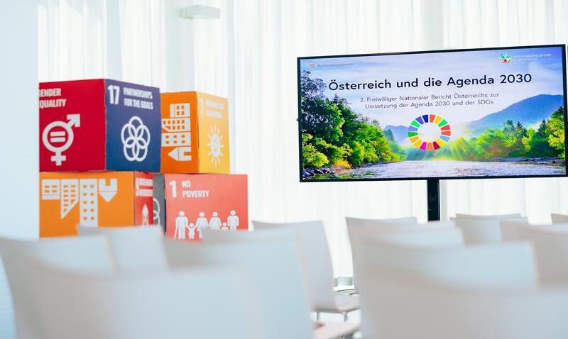 Ein Konferenzraum mit einem Bildschirm, der die Präsentation 'Österreich und die Agenda 2030' zeigt. Im Vordergrund stehen bunte Würfel mit Symbolen und Nummern, die verschiedene Ziele der nachhaltigen Entwicklung (SDGs) repräsentieren, wie Geschlech