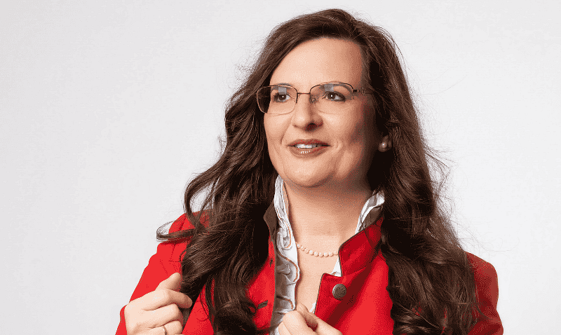 Frau mit langen, welligen Haaren in roter Jacke über weißem Shirt, hält die Jackenrevers mit beiden Händen. Sie trägt im Gesicht eine Brille