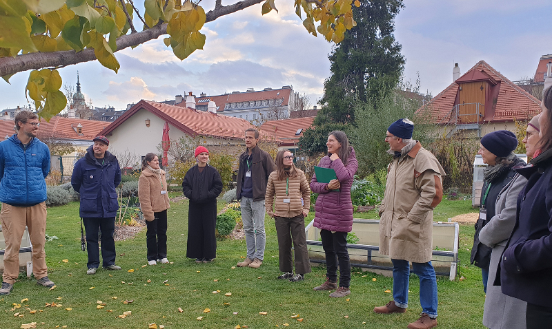 Eine Gruppe von Menschen steht in einem Garten, einige tragen warme Kleidung. Im Hintergrund sind Häuser mit roten Dächern zu sehen. Der Himmel ist bewölkt und ein Baum mit gelben Blättern ragt ins Bild.
